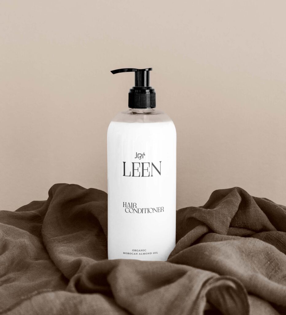 Minimalist white LEEN hair conditioner bottle with black pump dispenser, displayed on soft brown fabric against a neutral beige background.